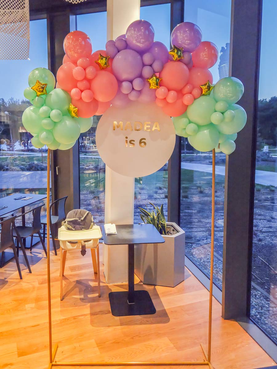 Dekoracija za dječji rođendan s pastelno obojenim balonima (ružičasti, ljubičasti i zeleni) raspoređenima u luk oko okrugle ploče s natpisom „MADEA is 6“. Između balona nalaze se mali zlatni baloni u obliku zvijezda.